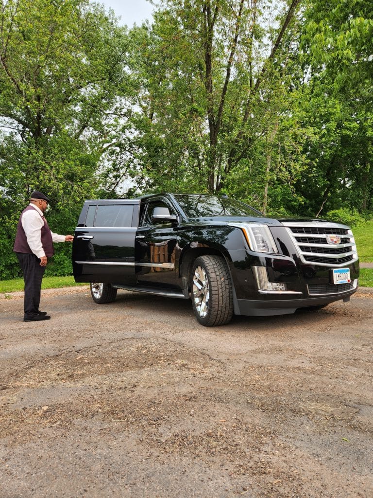 Hourly Driver Minneapolis. CMB Transport chauffeur opening door of luxury black Cadillac Escalade, showcasing professional VIP transportation service in Minneapolis and Twin Cities.
